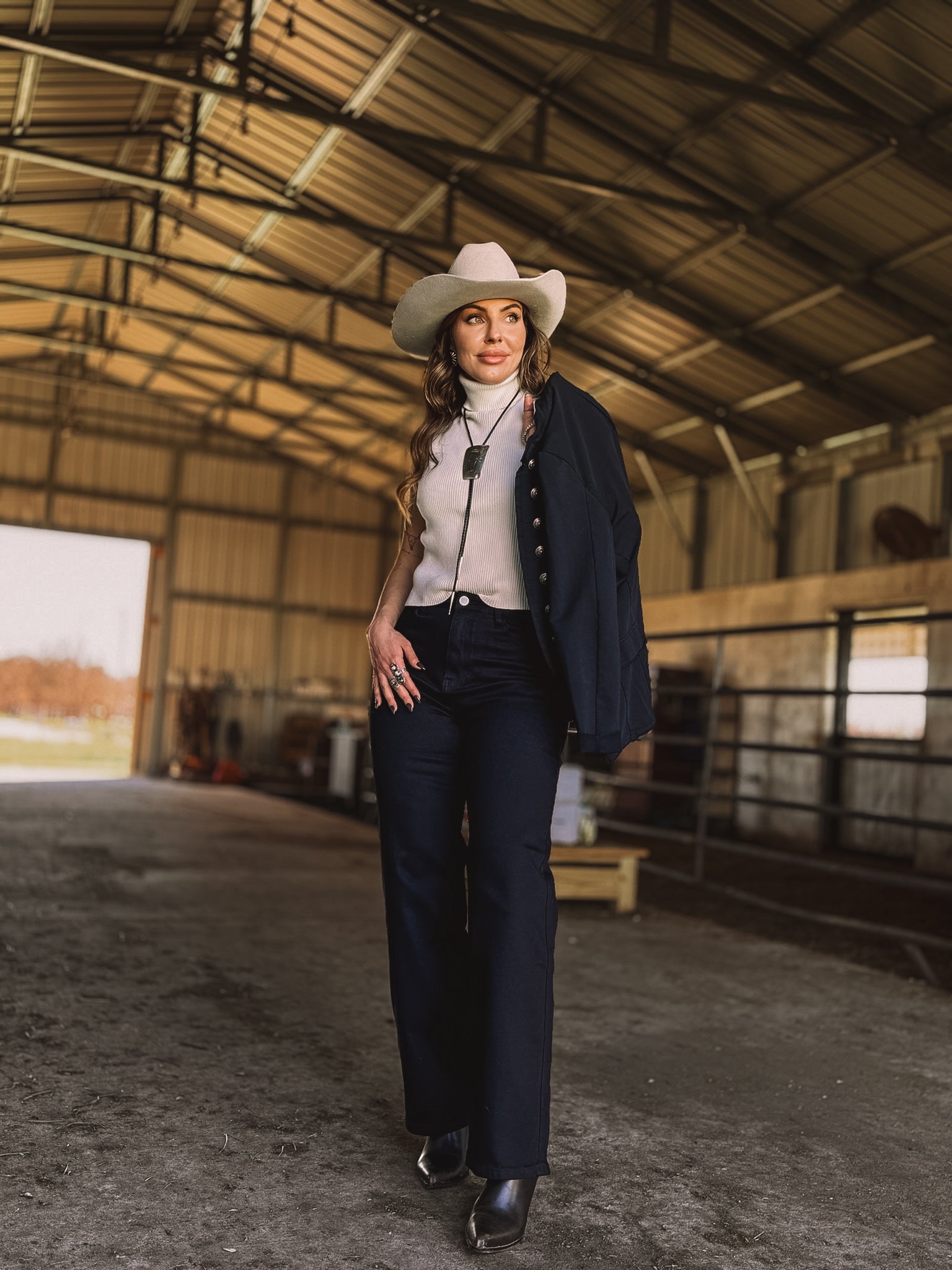 Woman in a white cowboy hat and western boho outfit walking in a barn, NFR style fashion.