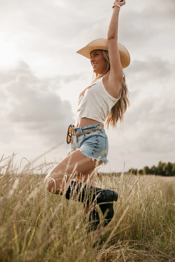 Woman in a cowboy hat and denim shorts standing in a field with a cloudy sky.