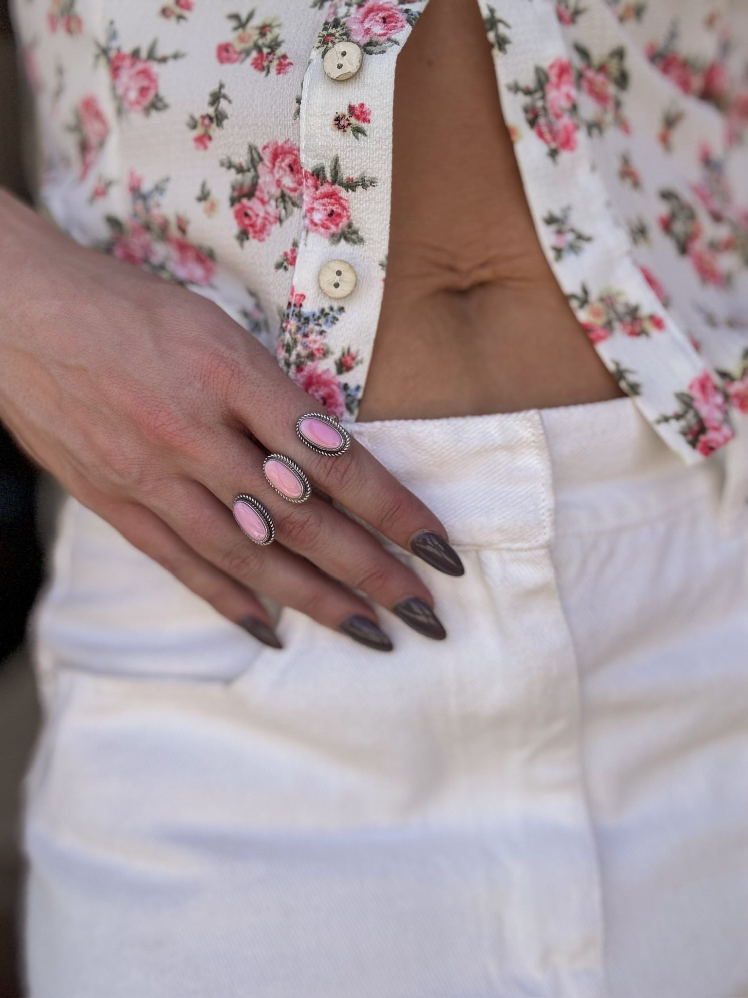 Woman wearing floral blouse, white jeans, pink stone western rings, and dark almond nails