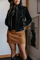 Woman wearing western boho outfit with black jacket, tan mini skirt, black cowgirl boots and hat