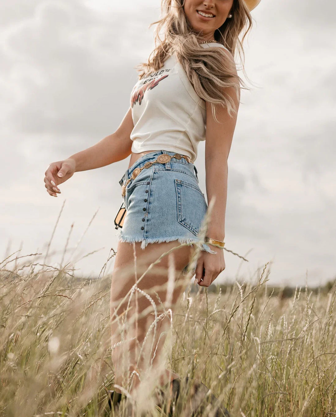Woman in a white crop top and denim shorts standing in a field with a cloudy sky.