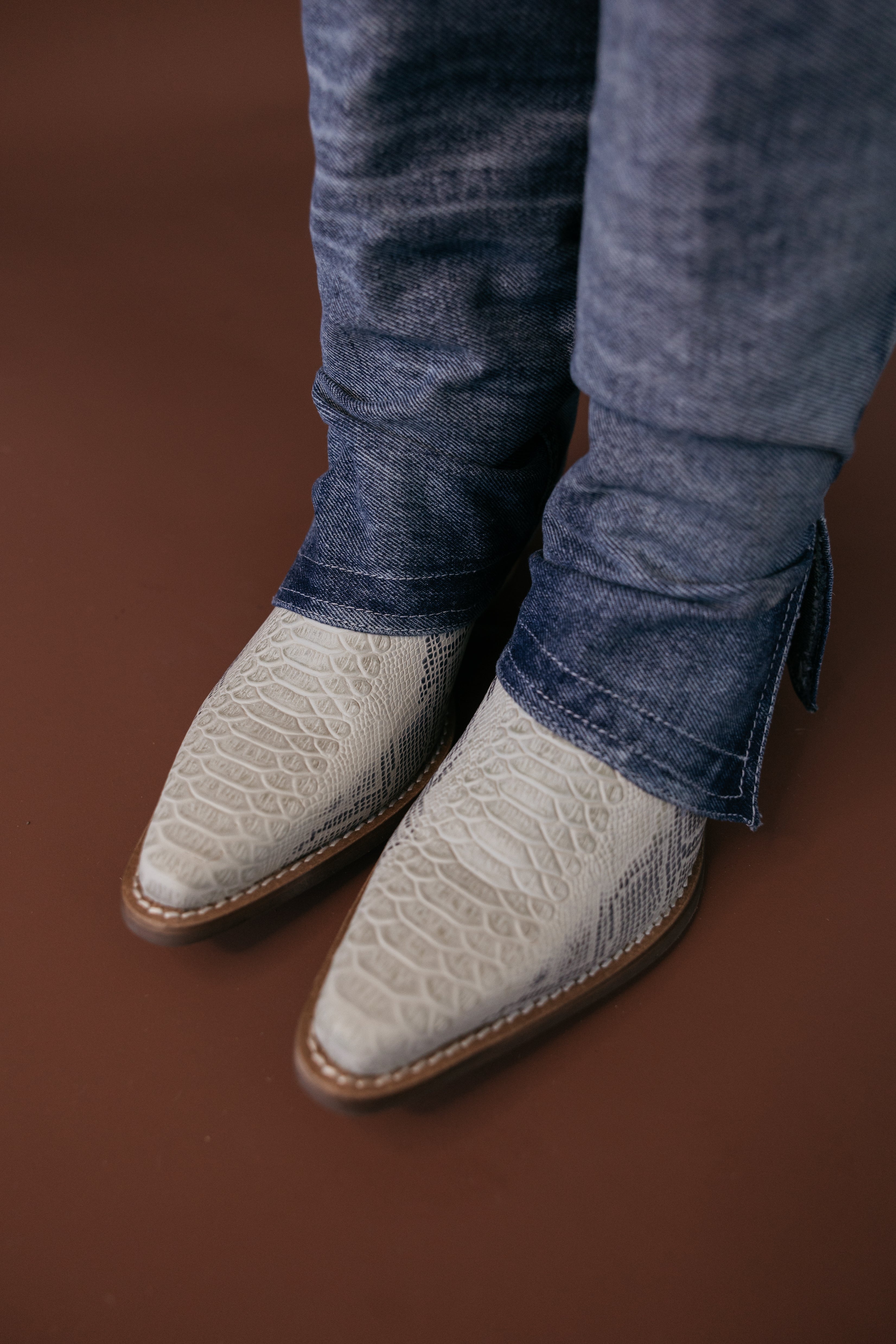 White textured western cowboy boots under blue jeans on a brown background