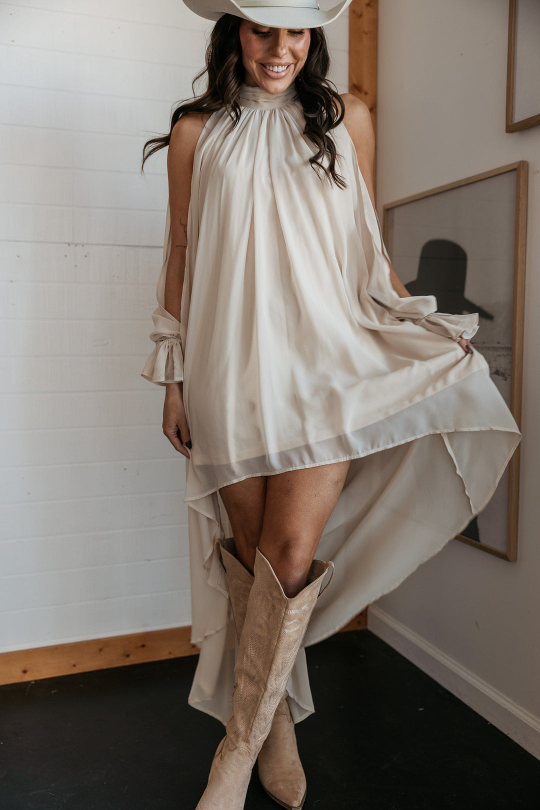 Woman in a white dress and beige boots standing indoors with a white wall and framed picture in the background.