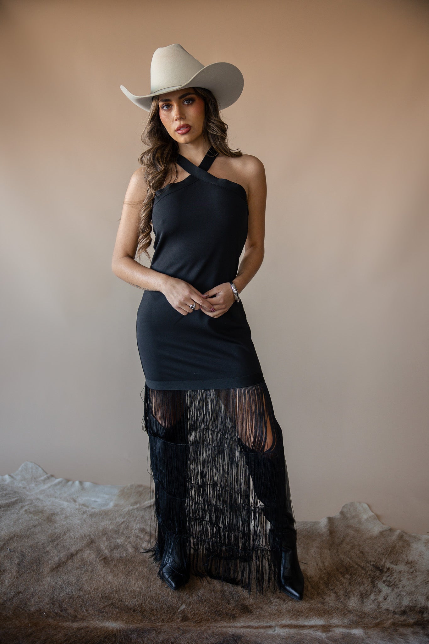 Woman in a black western fringe dress, cowboy hat, and boots, standing on a cowhide rug.