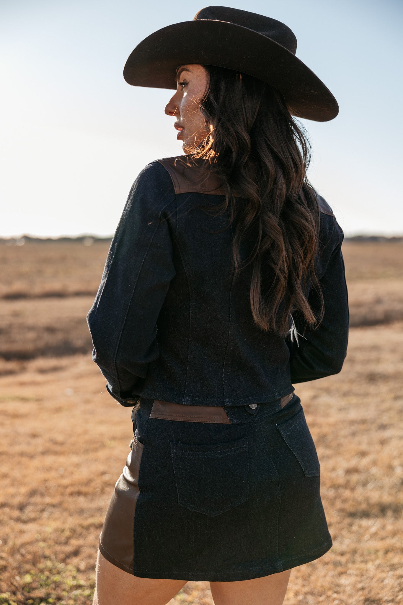 Woman in western denim jacket, mini skirt, and cowboy hat standing outdoors in a field