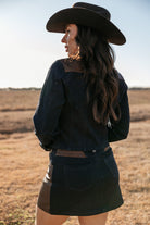 Woman in western denim jacket, mini skirt, and cowboy hat standing outdoors in a field