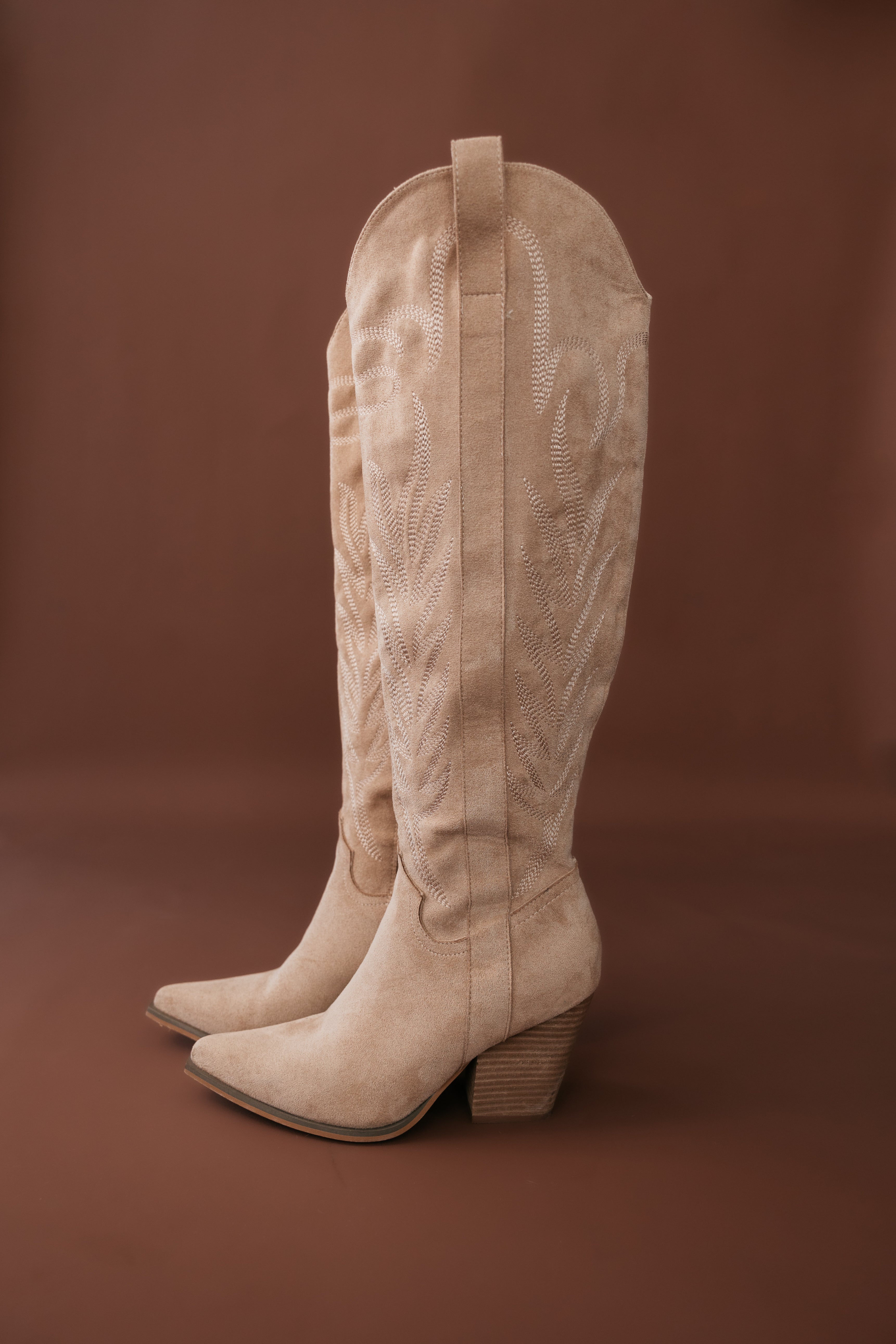 Tan suede western cowgirl boots with embroidery and block heels on brown background