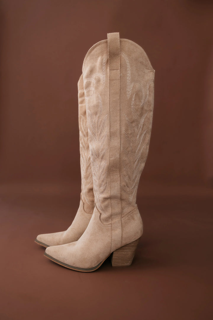 Tan suede western cowgirl boots with embroidery and block heels on brown background
