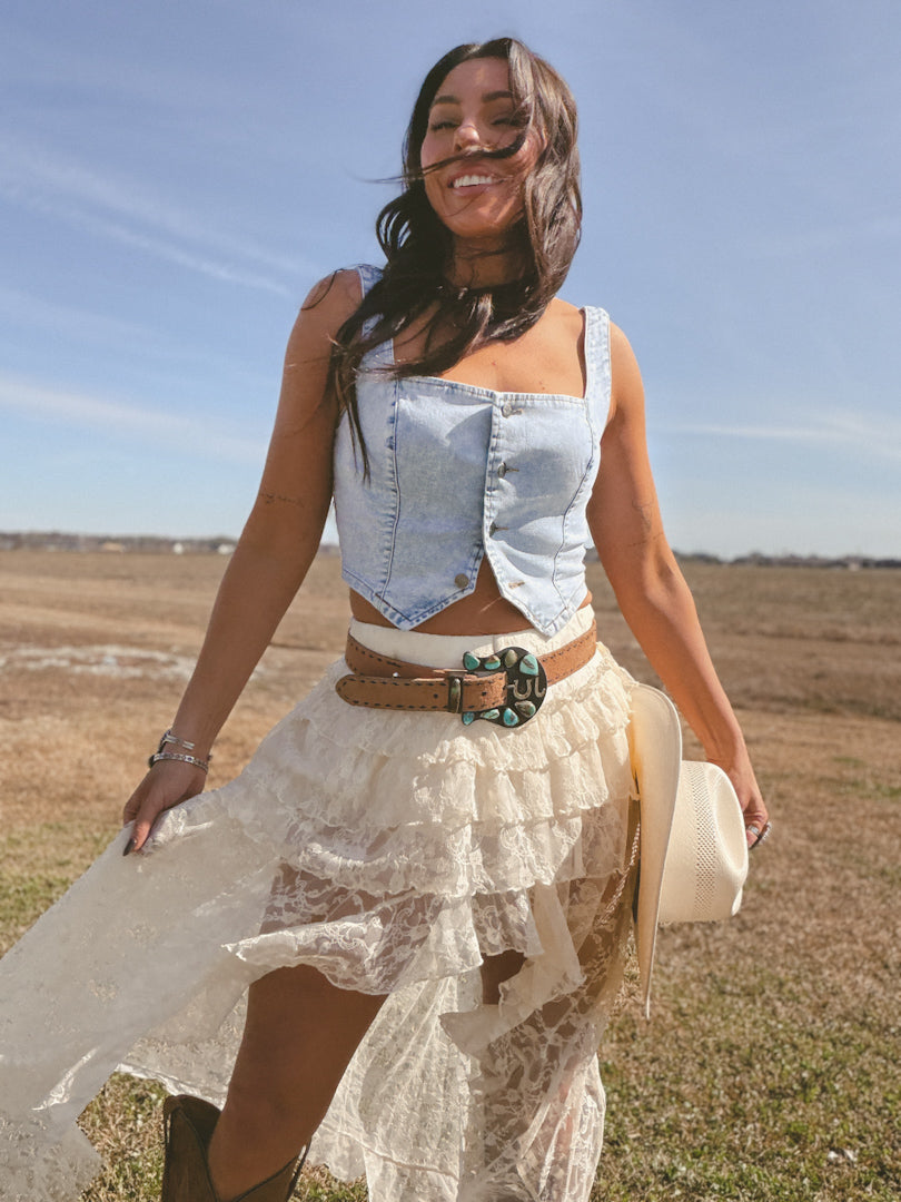Woman in denim vest and lace skirt with western belt, holding cowboy hat outdoors