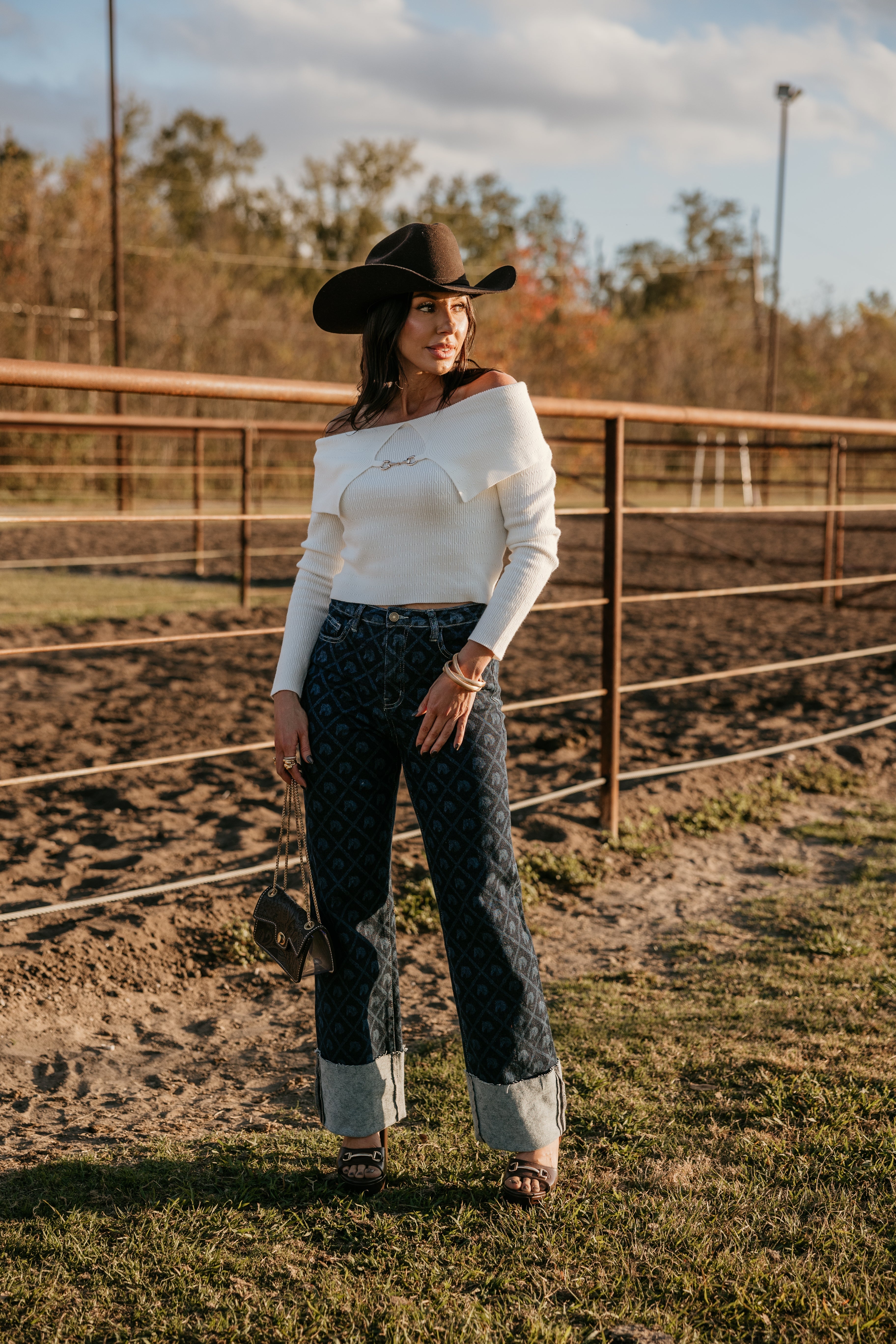 Woman in a cowboy hat, off-the-shoulder sweater, patterned wide-leg jeans, and heels at a ranch.