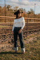 Woman in a cowboy hat, off-the-shoulder sweater, patterned wide-leg jeans, and heels at a ranch.