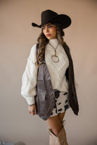 Woman in cowgirl hat, white sweater, brown vest, cow print skirt, and cowboy boots