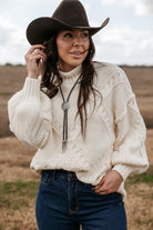 Woman in a brown cowboy hat, cream knit sweater, bolo tie, and jeans in a western field