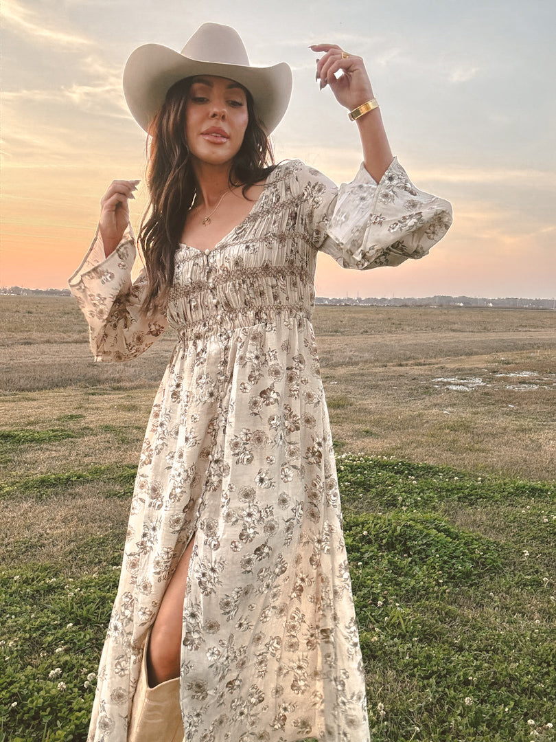 Woman in a floral dress and cowboy hat standing in a field with a sunset.