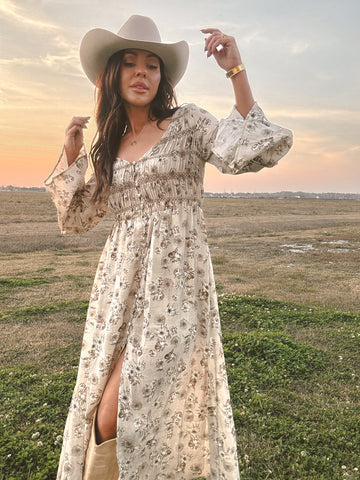 Woman in a floral dress and cowboy hat standing in a field with a sunset.