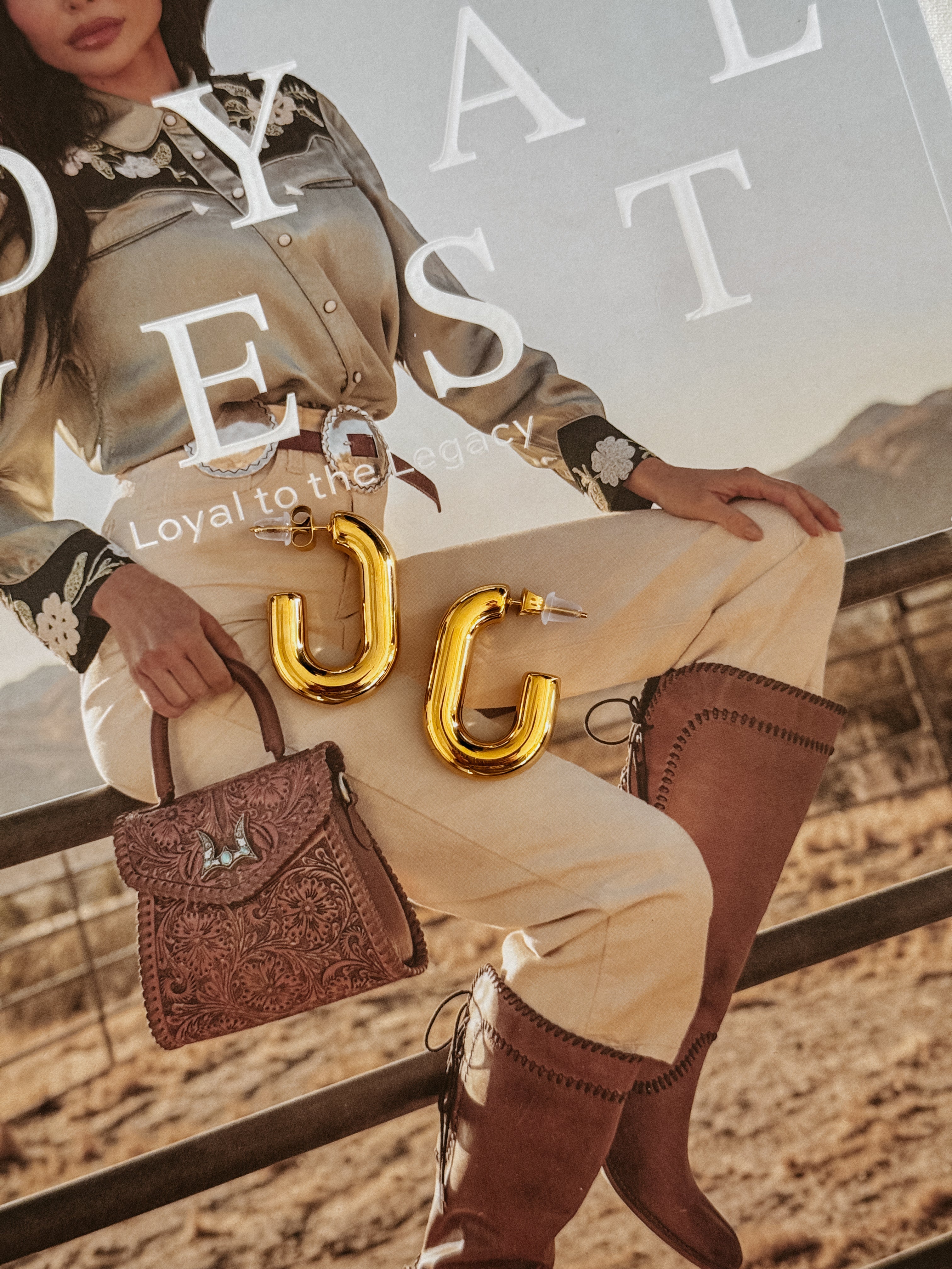 Gold western-style hoop earrings on magazine featuring cowgirl in embroidered shirt, boots, tooled purse.