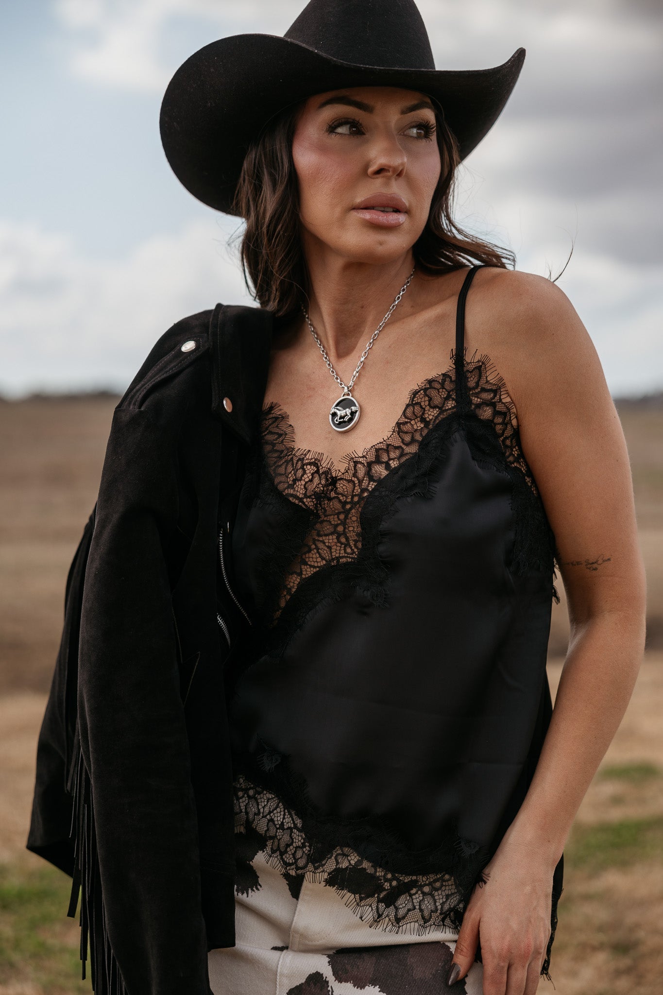 Woman in a black cowboy hat, black lace camisole, suede fringe jacket, and cow print pants outdoors