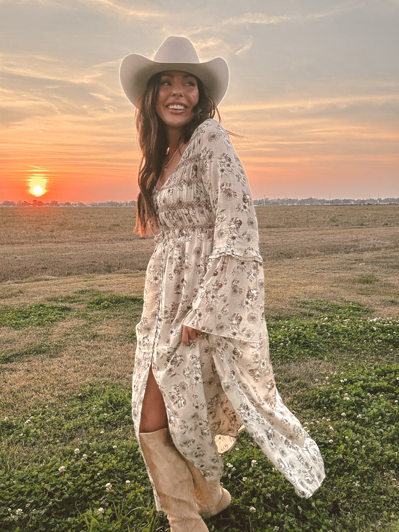 Woman in a floral dress and cowboy hat standing in a field at sunset