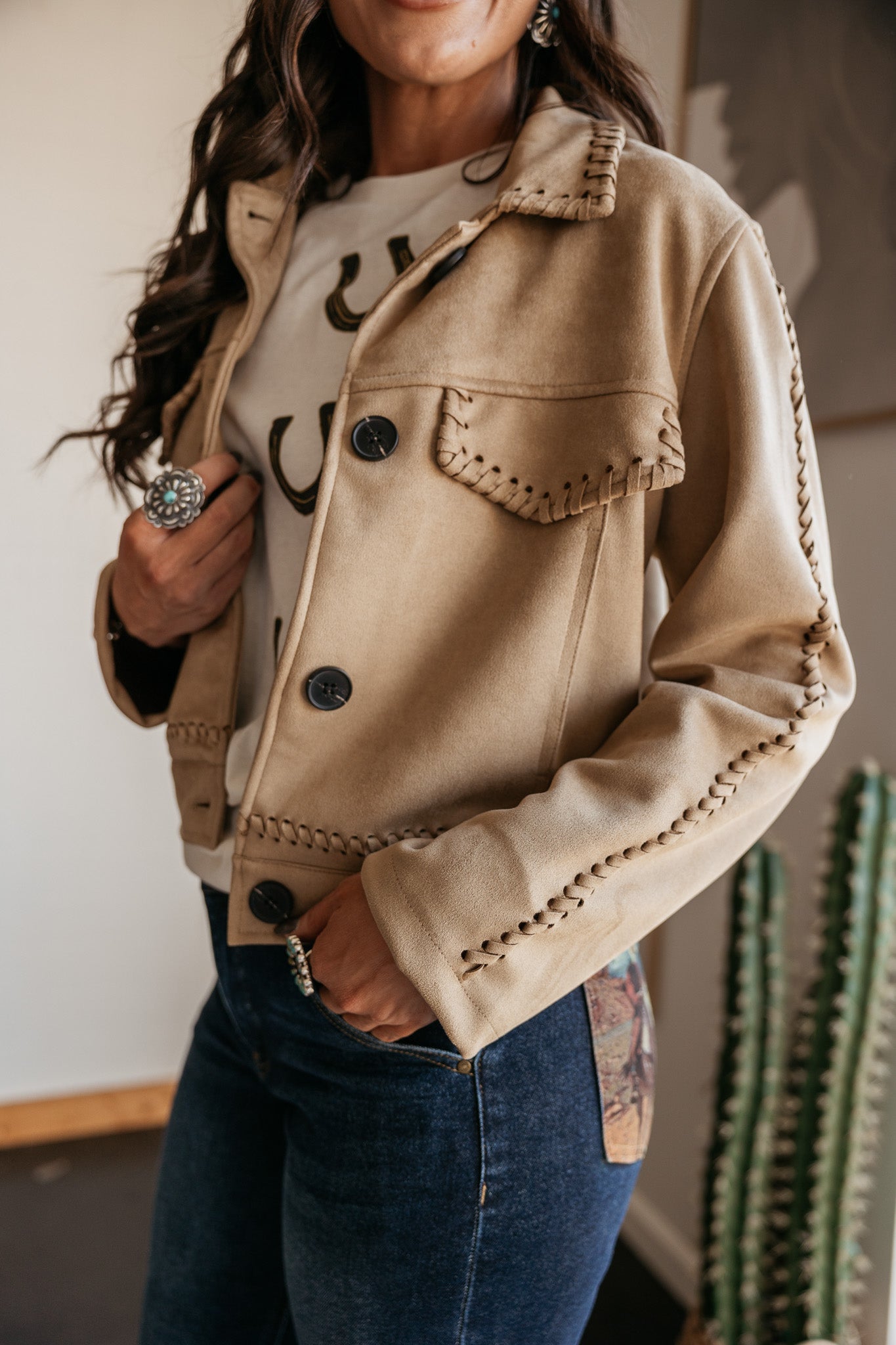 Person wearing a beige jacket with decorative buttons and trim, standing indoors with a cactus in the background.