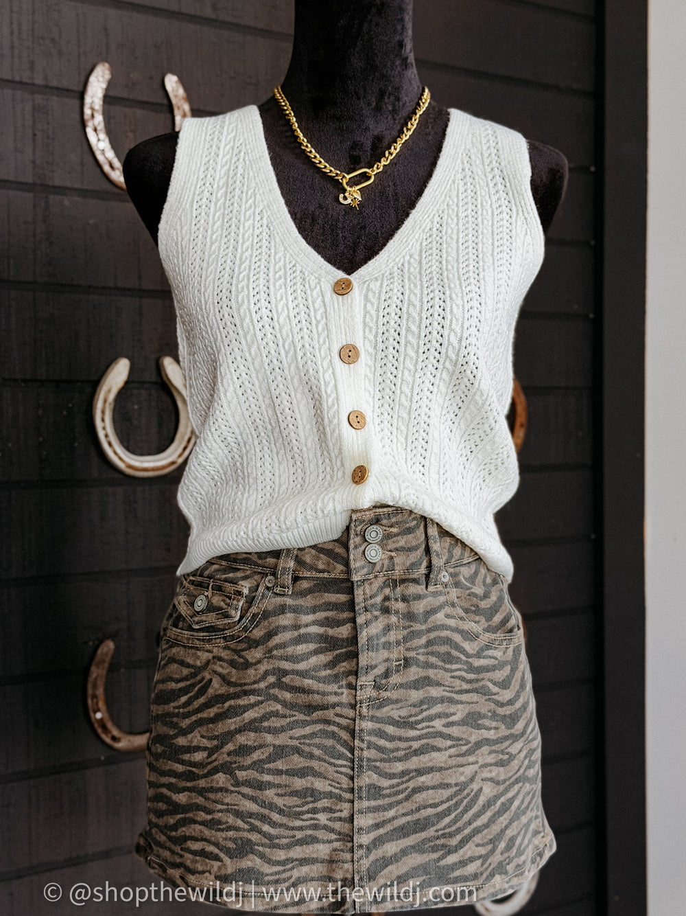 Mannequin wearing a white knitted vest and zebra print skirt with gold jewelry against a dark background.