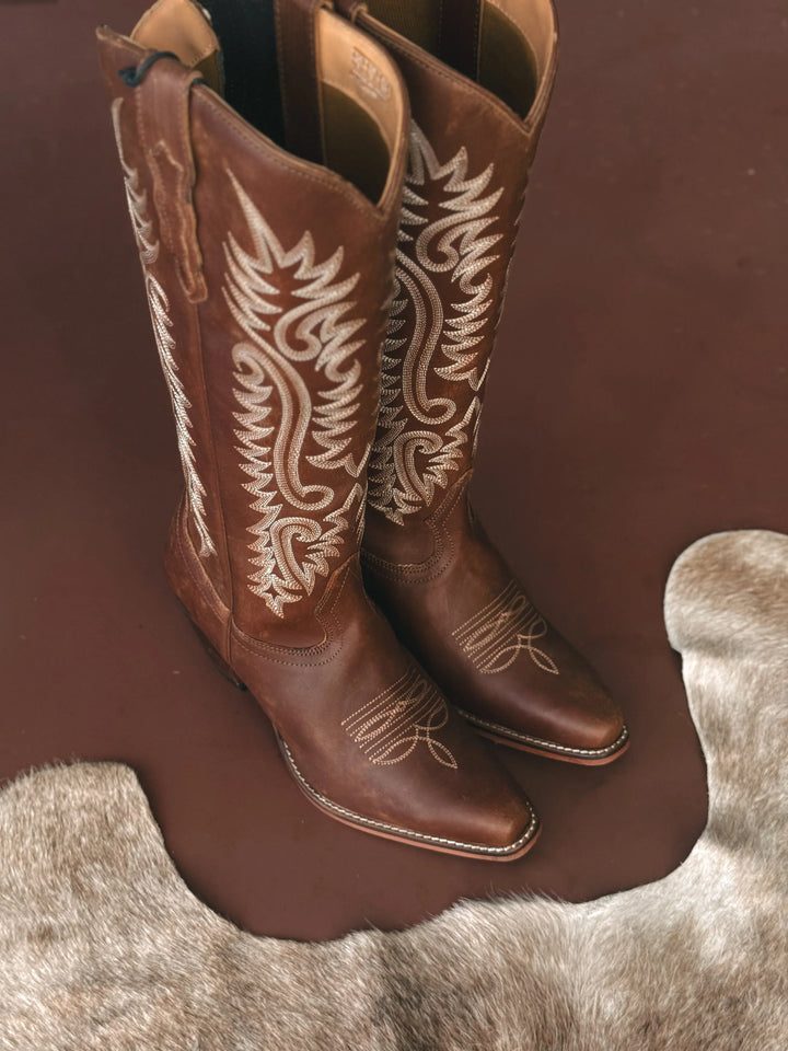 Pair of brown cowboy boots with white embroidery on a brown surface.
