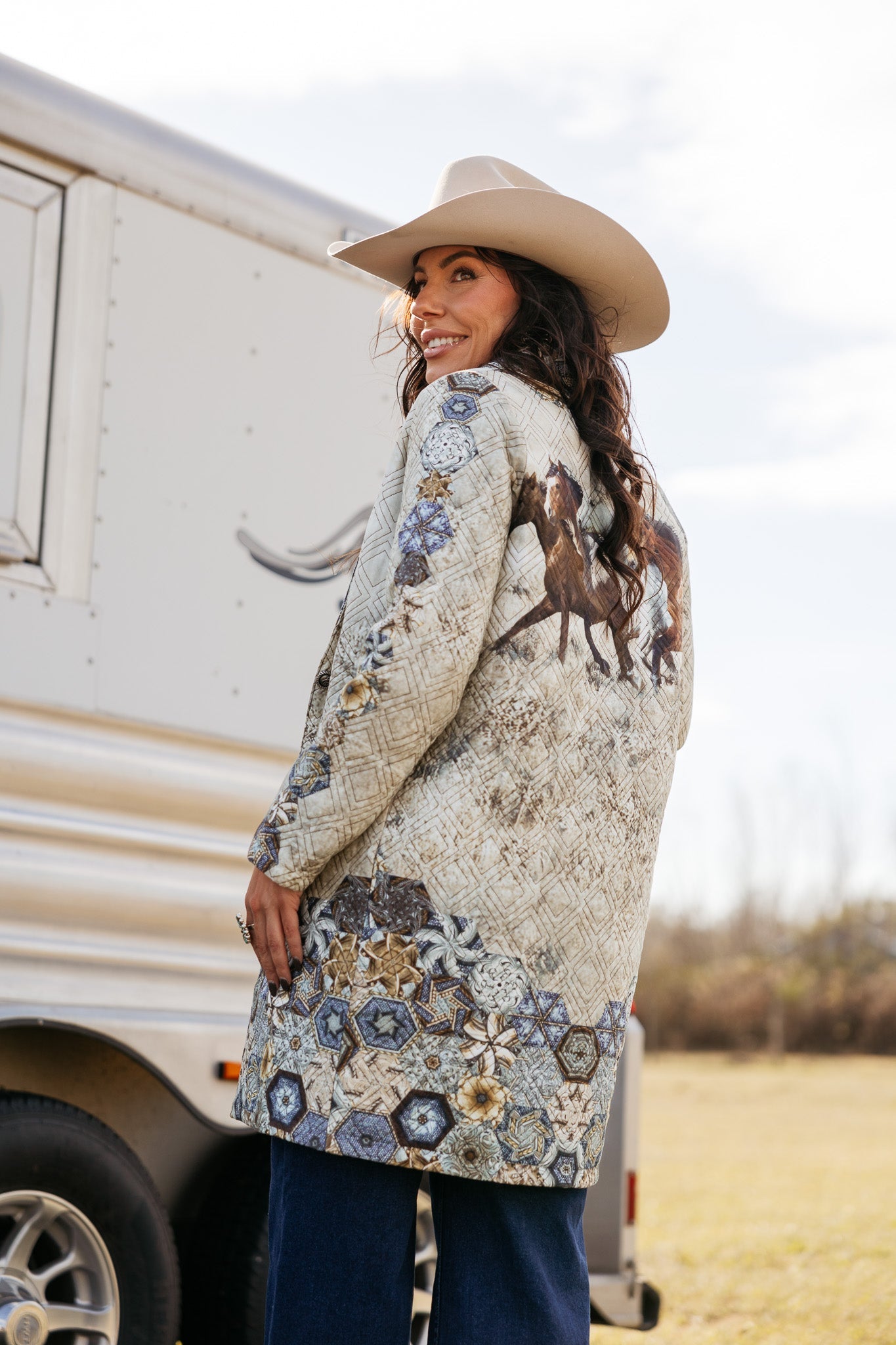 Woman in cowboy hat and boho western coat with horse print, standing by trailer outdoors