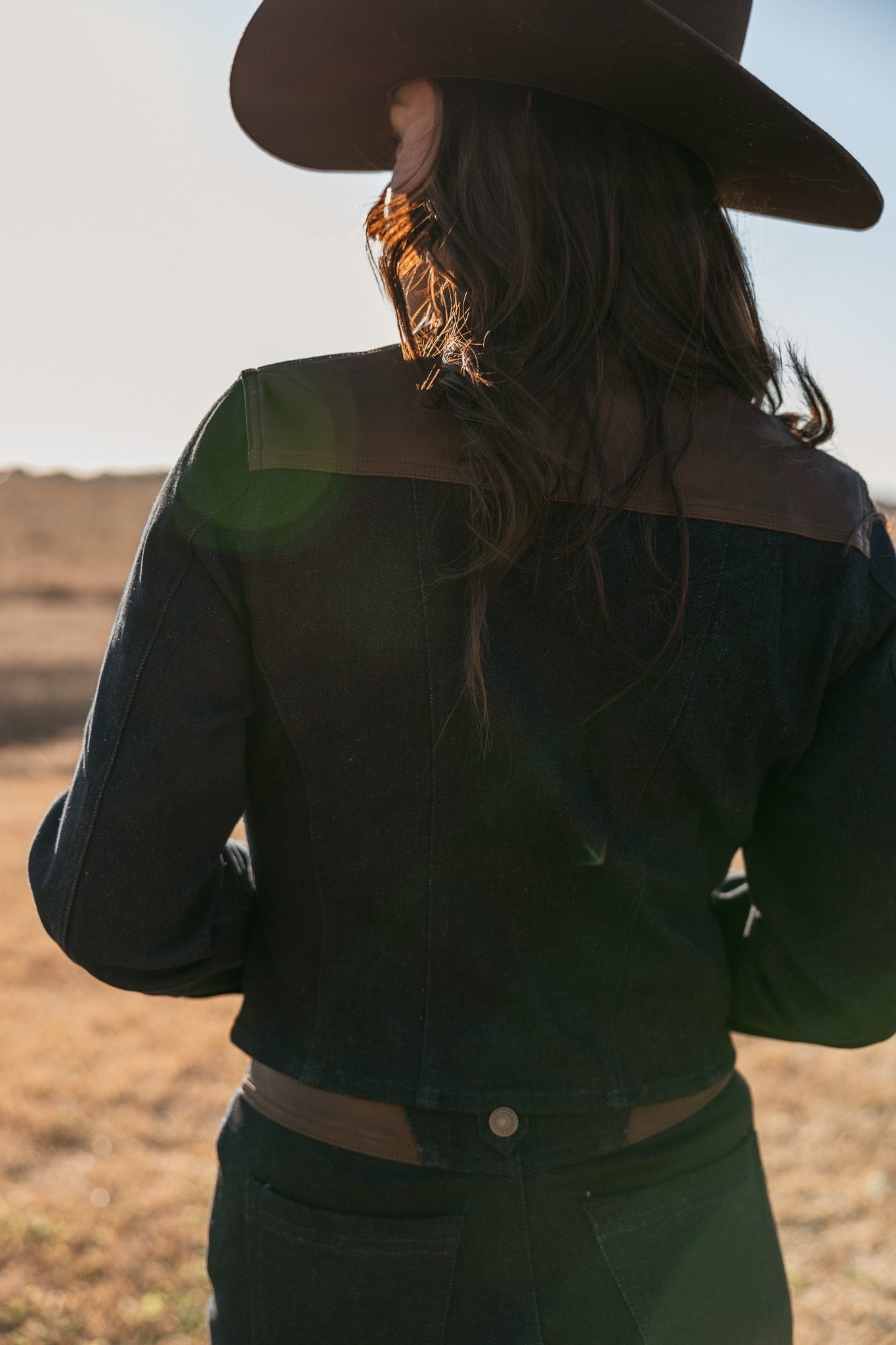 Woman in dark denim western jacket and cowboy hat, outdoors in sunlight, boho cowgirl style