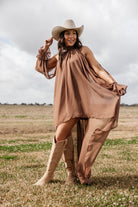 Woman in brown flowy western dress, ivory cowboy hat, and suede boots standing in a field
