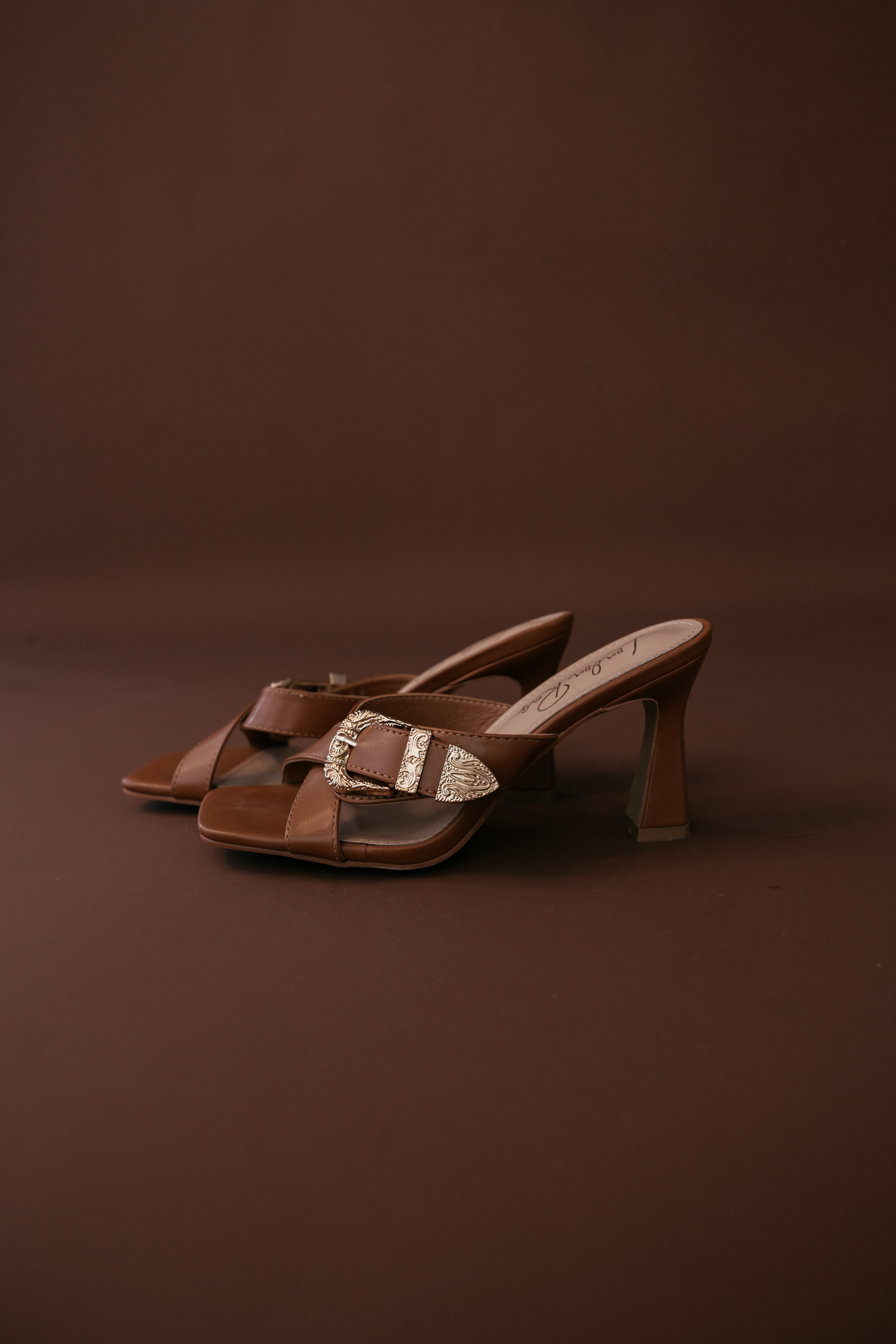 Brown western heeled sandals with gold buckle detail on brown background