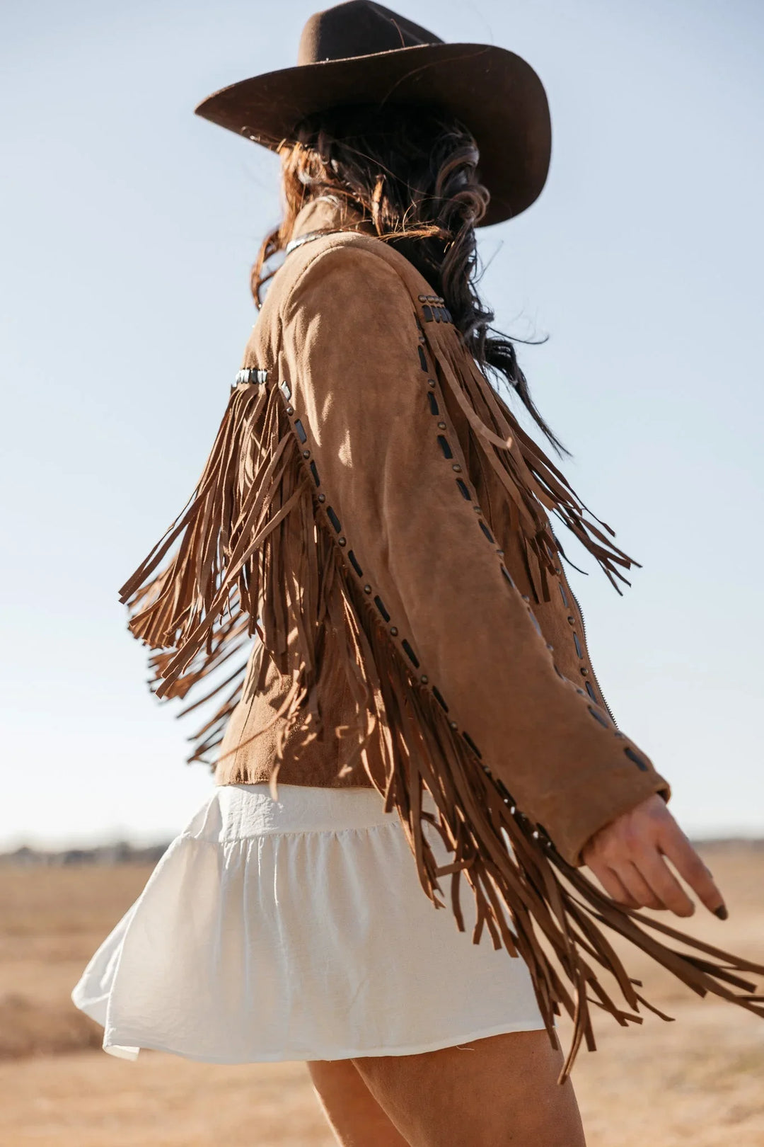 Woman in brown suede fringe jacket, white skirt, and western hat in a sunny outdoor field