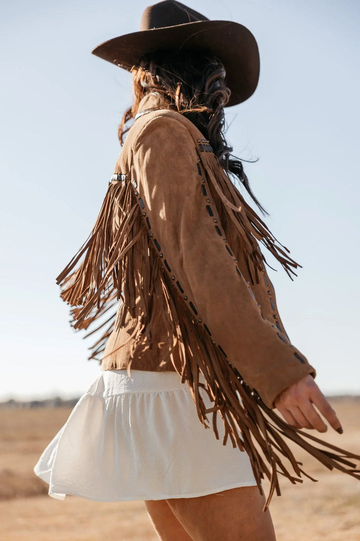 Woman in brown suede fringe jacket, white skirt, and western hat in a sunny outdoor field