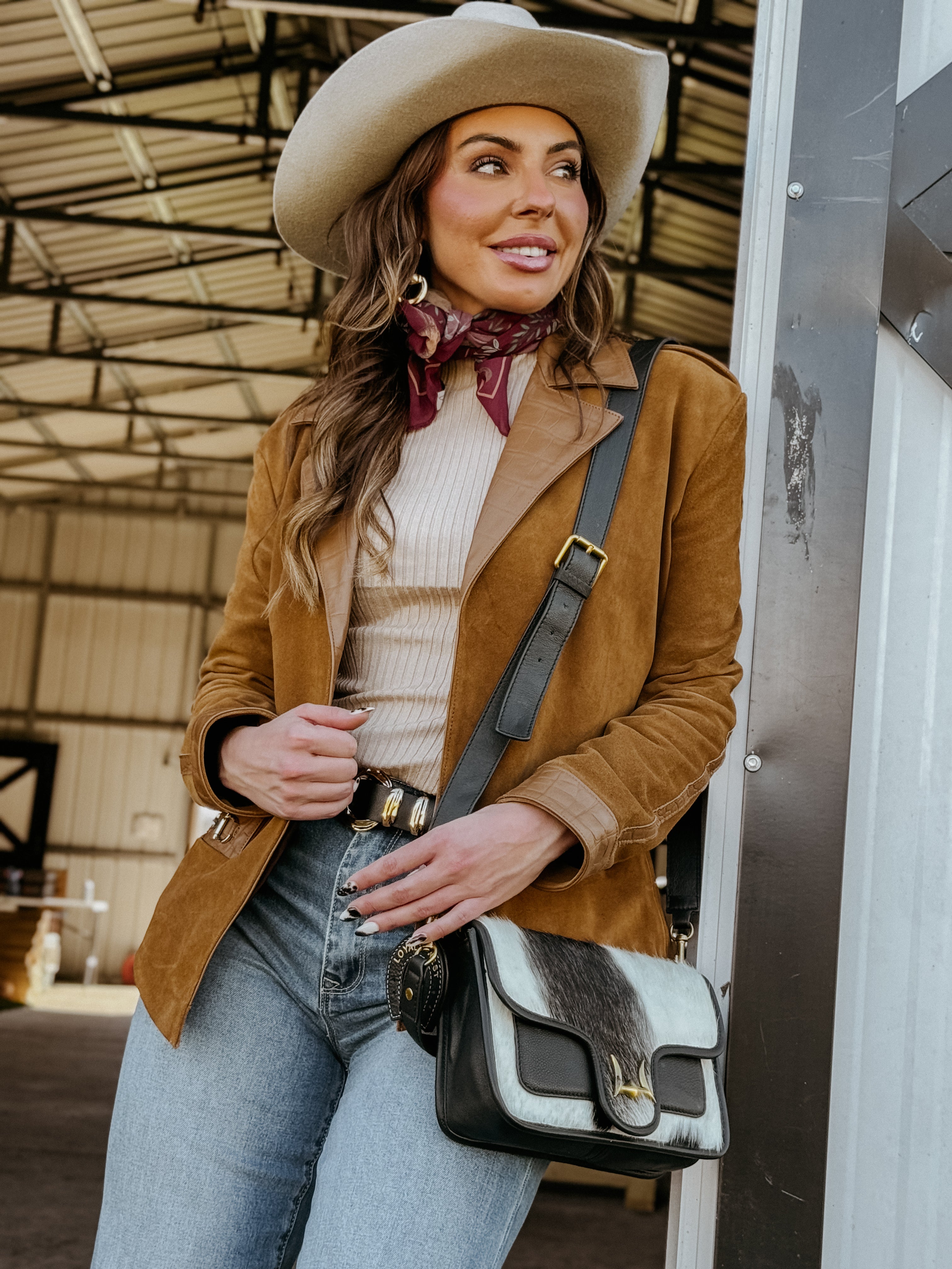 Woman in western boho outfit with tan suede jacket, wide-brim hat, and cow print bag