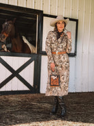 Woman in western boho dress, cowboy hat, and boots, standing by horse stable with horse.