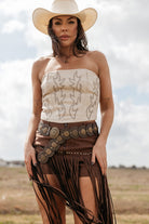 Woman in western boho outfit with white strapless top, fringed brown skirt, statement belt, and cowboy hat outdoors