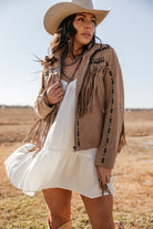 Woman in a tan fringe western jacket, white dress, and cowboy hat standing outdoors
