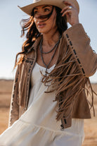 Woman in tan suede fringe jacket, white dress and cowboy hat, western boho style outdoors