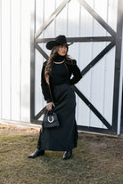 Woman in western boho outfit with black cowboy hat, long black skirt, boots, and horseshoe purse