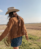 Woman in a tan fringe suede jacket, cowboy hat, and blue western dress standing outdoors