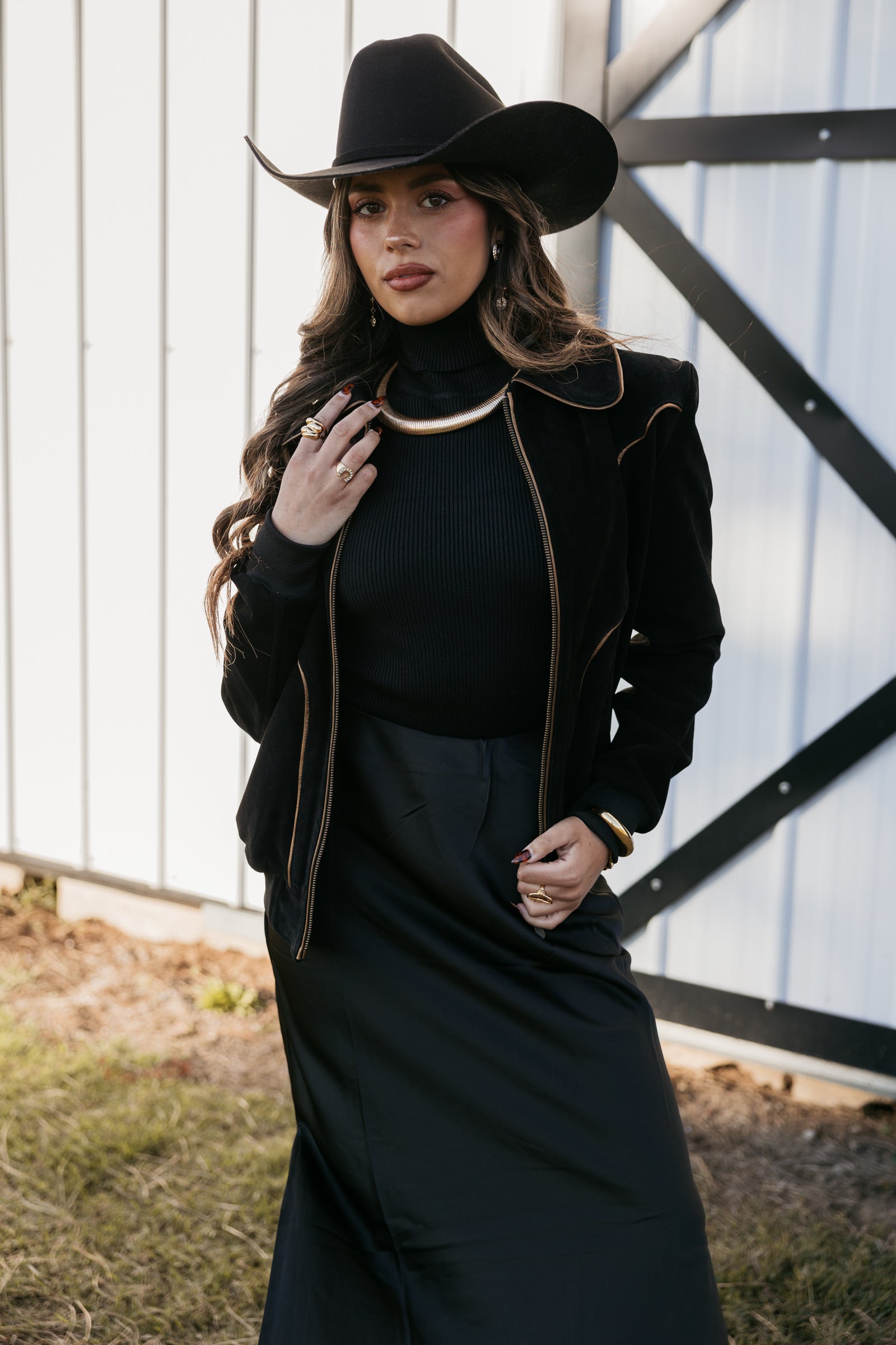 Woman in a black outfit with a black cowgirl hat standing in front of a white wall.