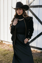 Woman in black western hat and boho outfit with gold jewelry, standing by barn door