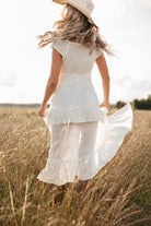 Woman in a white dress and hat standing in a field of tall grass.