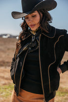 Woman in western black suede jacket, black cowboy hat, and scarf, outdoors in a field