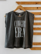Gray sleeveless shirt with 'Cowgirl Era' text on a wooden hanger against a wooden panel background