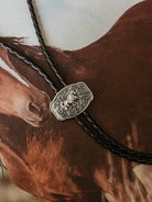 Western bolo tie with engraved horse concho on braided leather, styled on horse background.