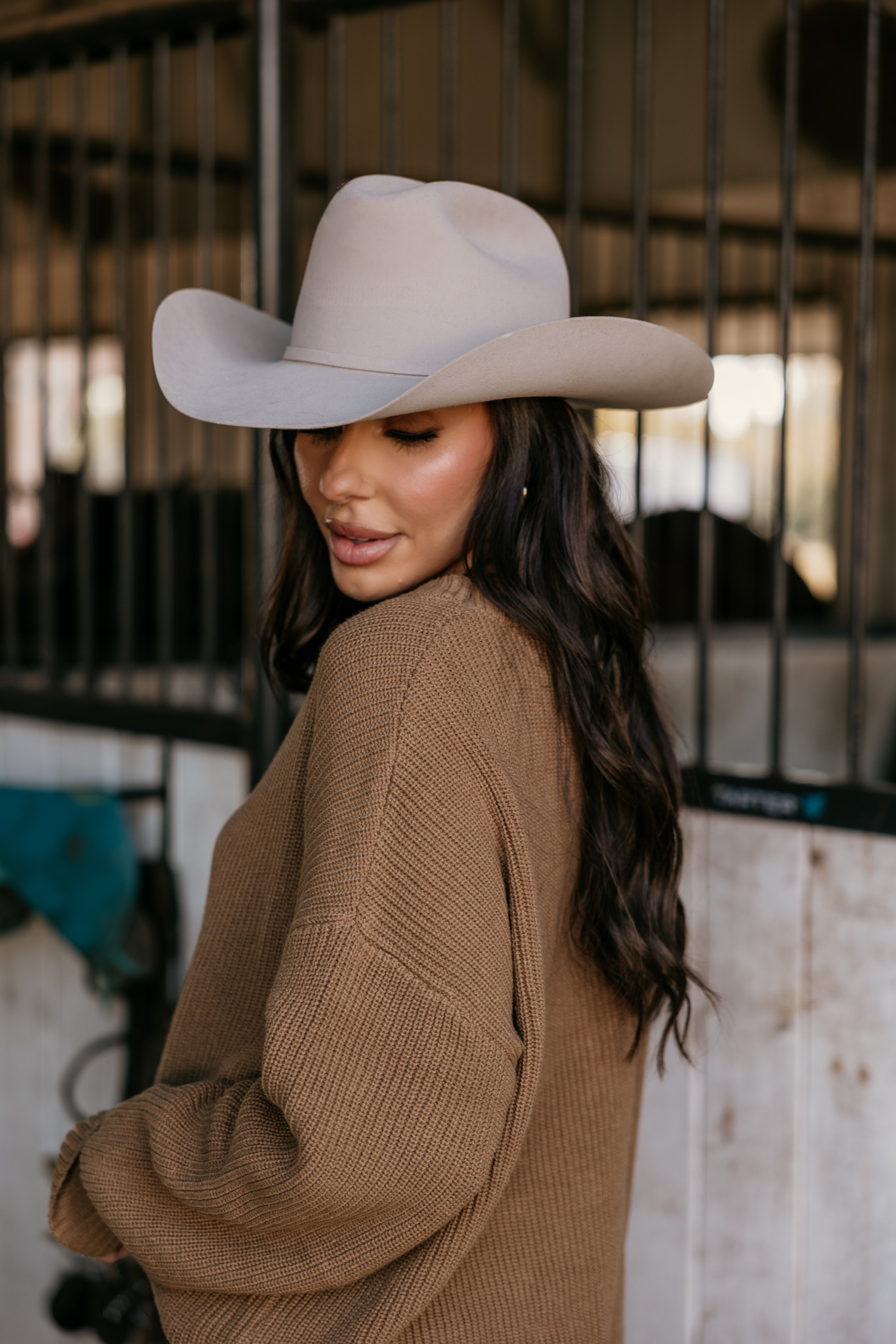 Woman in beige cowboy hat and brown sweater, western style, in front of horse stable