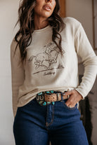 Woman wearing a cream western graphic sweatshirt with 'Saddle up' text, turquoise concho belt, and blue jeans