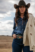 Woman in cowgirl hat and denim shirt wearing turquoise western belt, holding tan jacket, outdoors