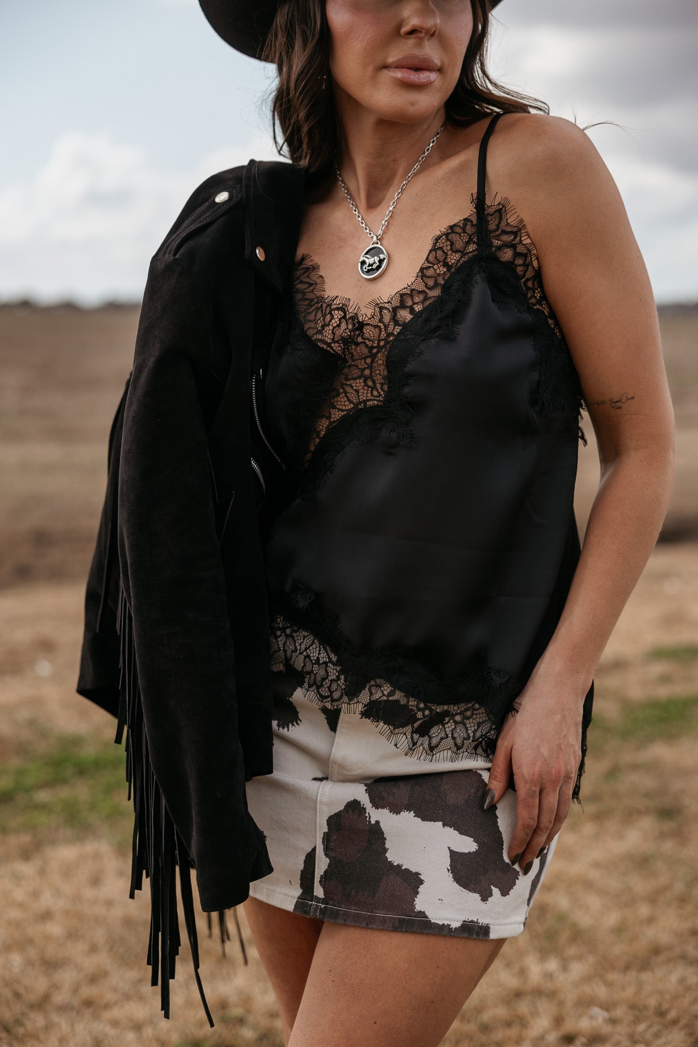 Woman in cow print skirt, black lace cami, black fringe jacket, and silver western necklace