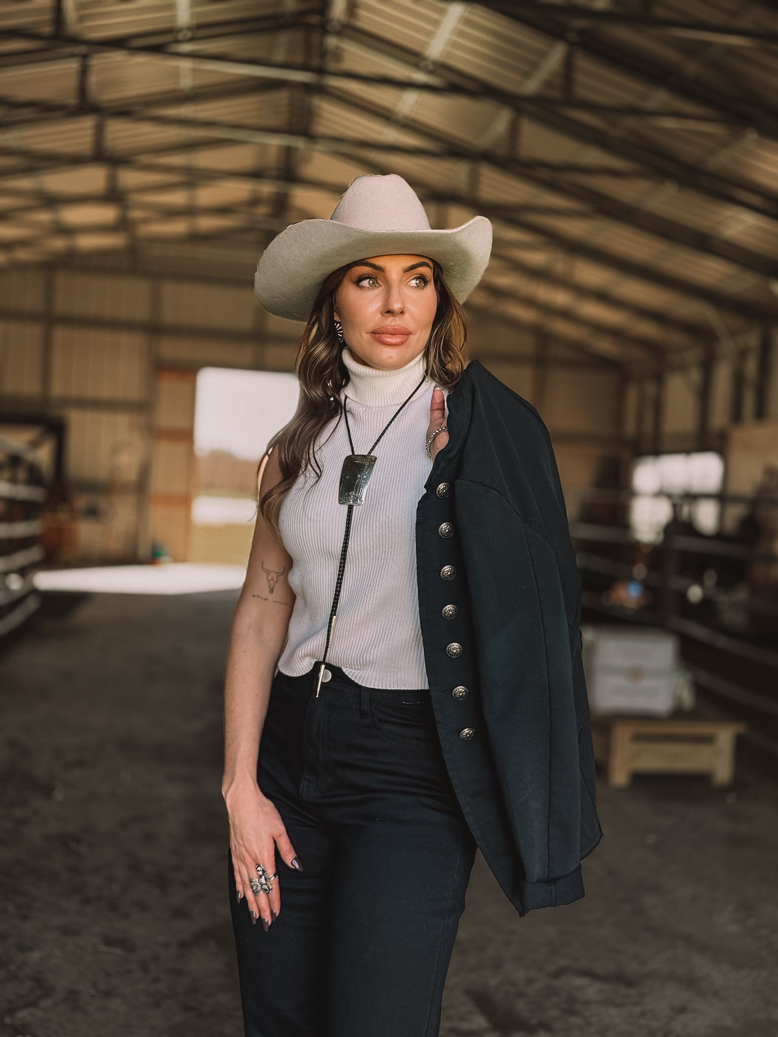 Woman in cowgirl hat, western necklace, white sleeveless top, black pants and jacket, standing in barn