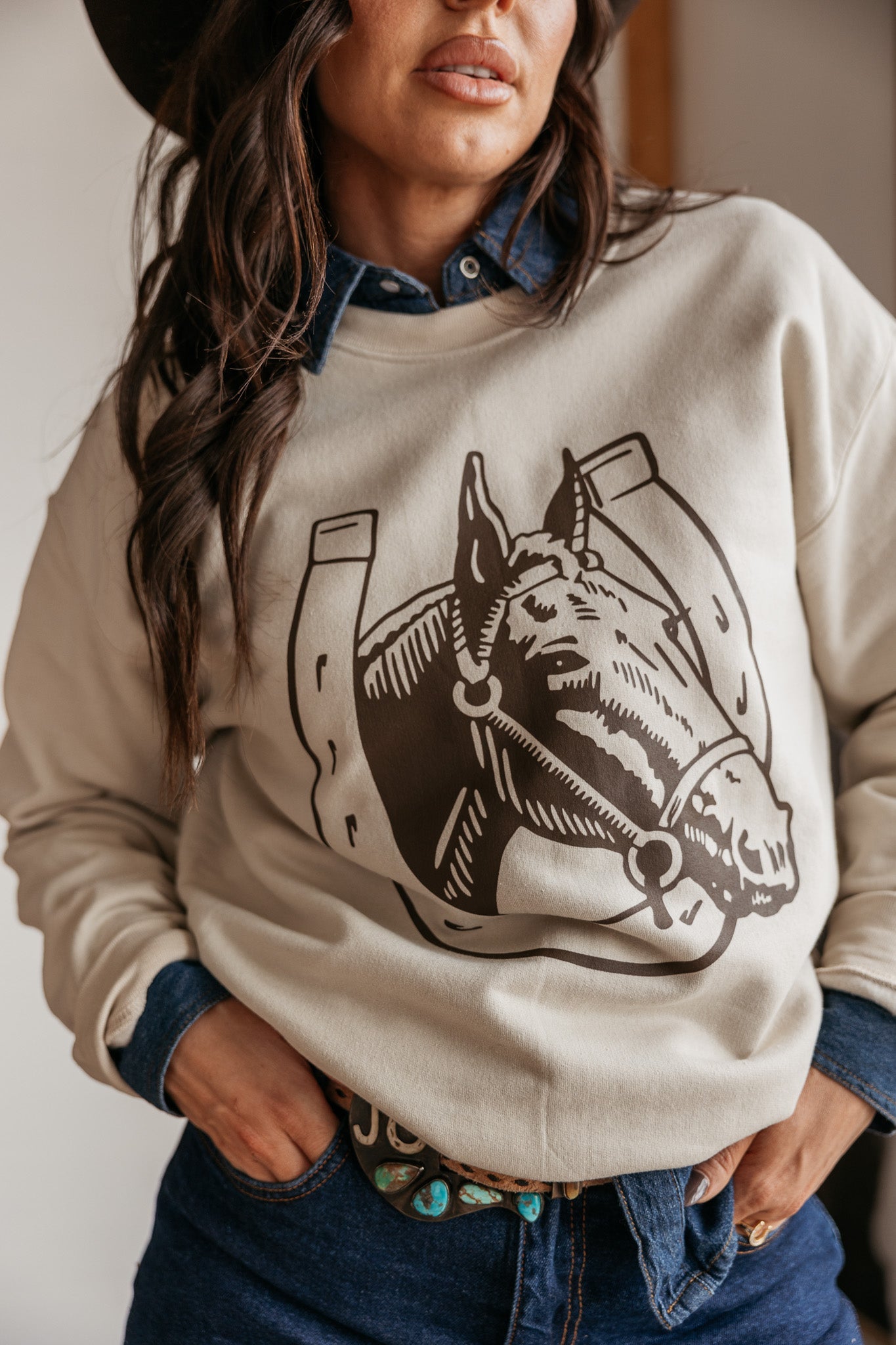 Woman in western graphic sweatshirt with horse and horseshoe, denim shirt, and turquoise belt
